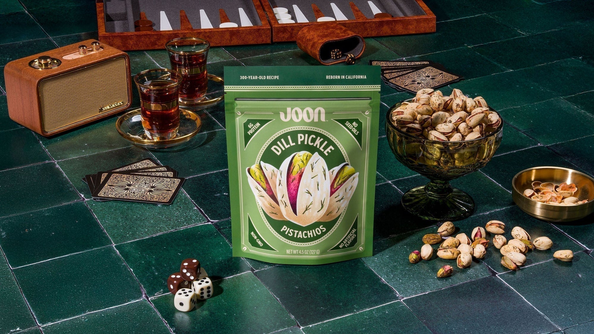 Joon Dill Pickle Pistachios bag surrounded by pistachios on a green tabletop.