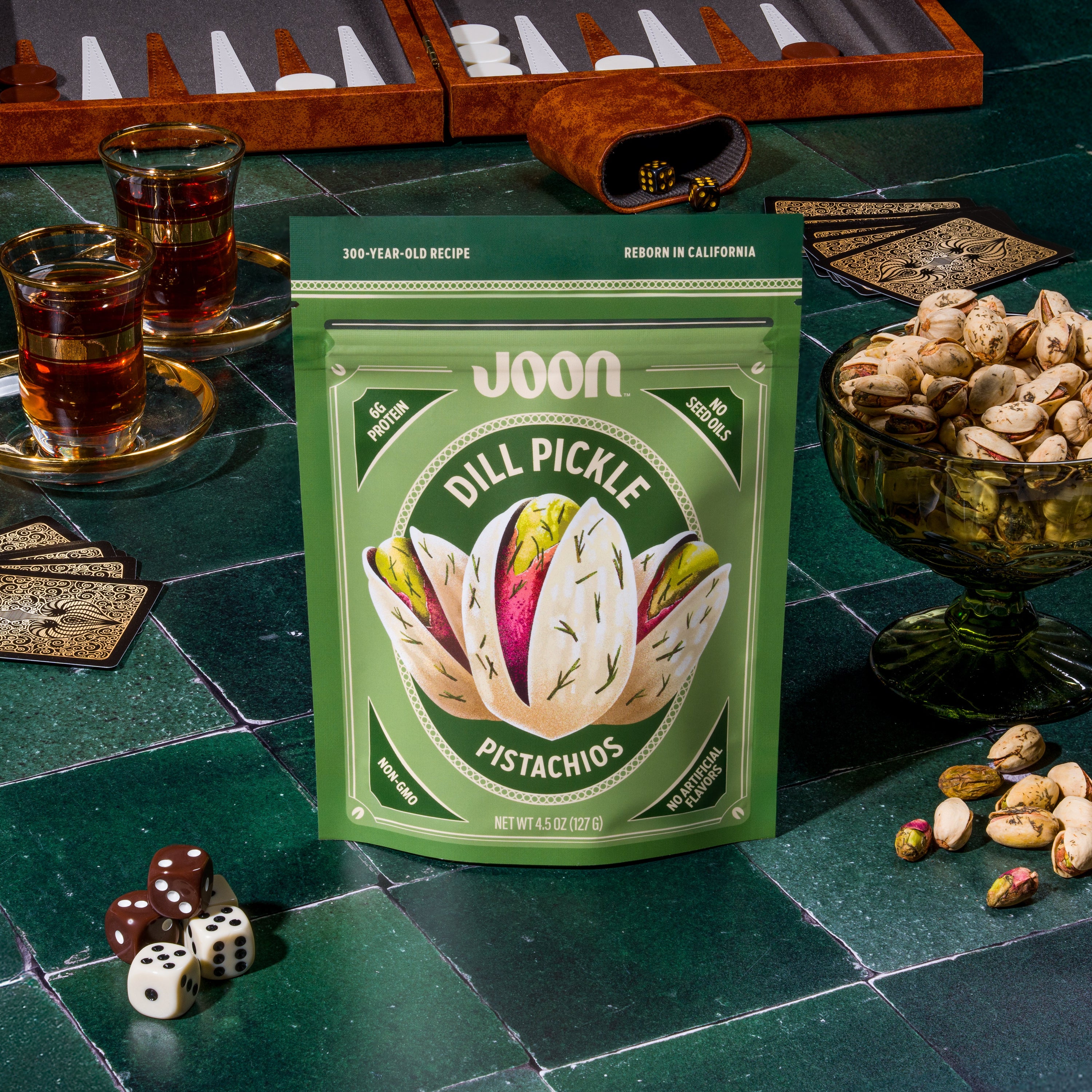 Joon Dill Pickle Pistachios 4.5 oz bag on a green game table with backgammon, cards, dice, tea, and pistachios.