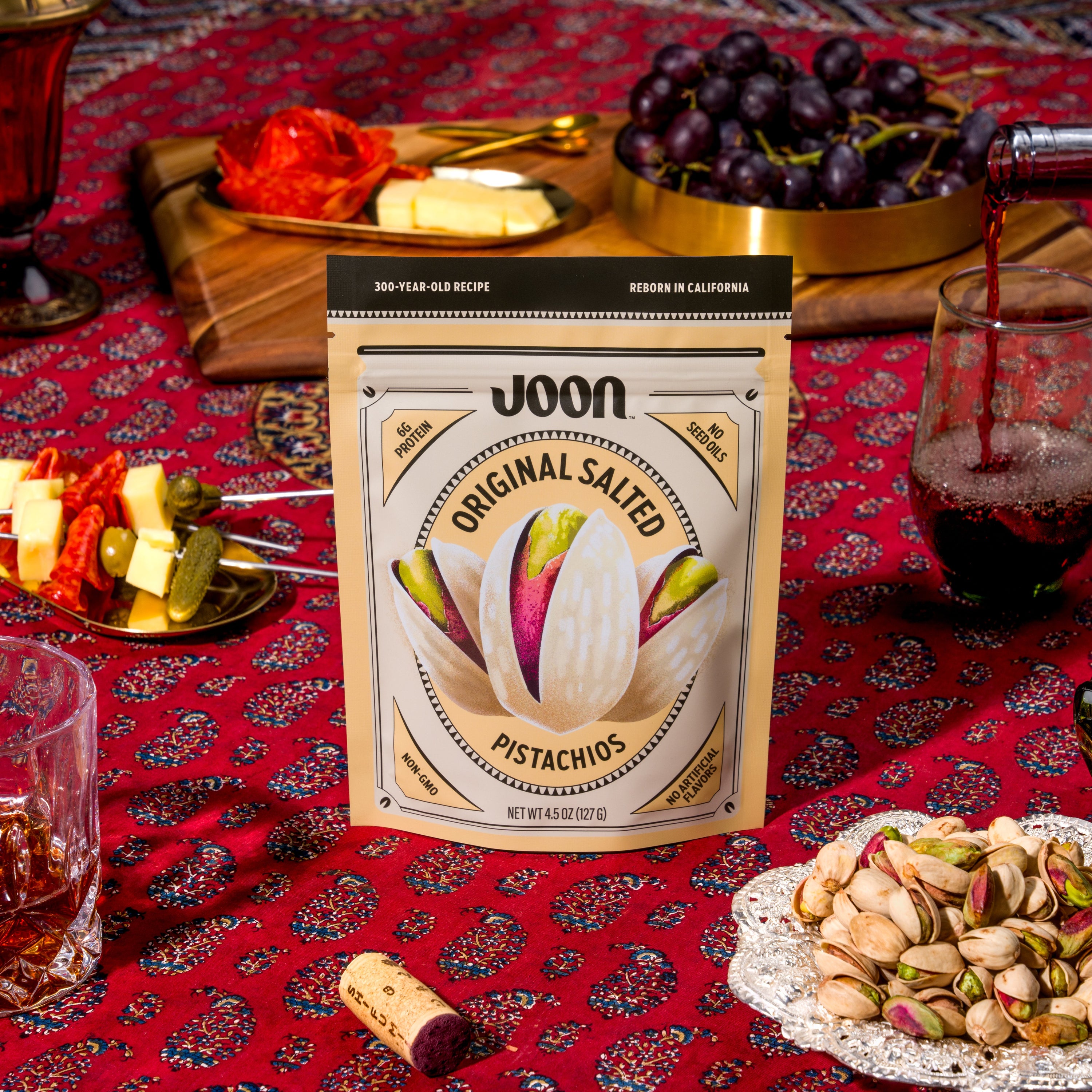 Joon Original Salted Pistachios 4.5 oz bag on a red table with grapes, cheese, wine, and pistachios.