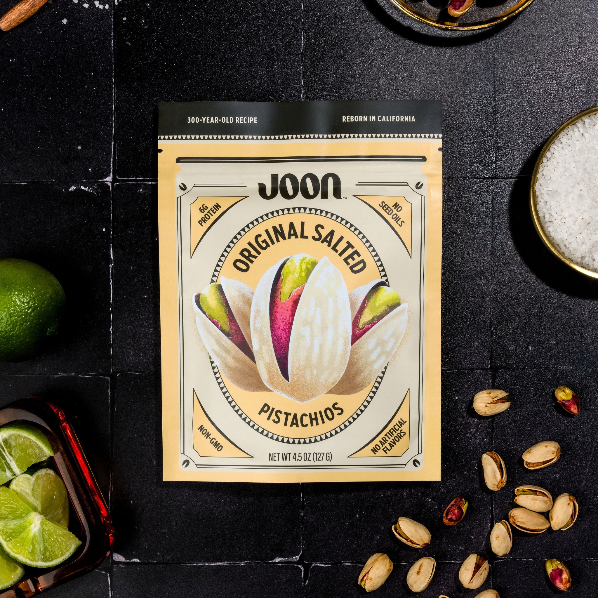 Packaging of Joon's Original Salted Pistachios with the product visible in the background, alongside limes and loose pistachios scattered on the surface.