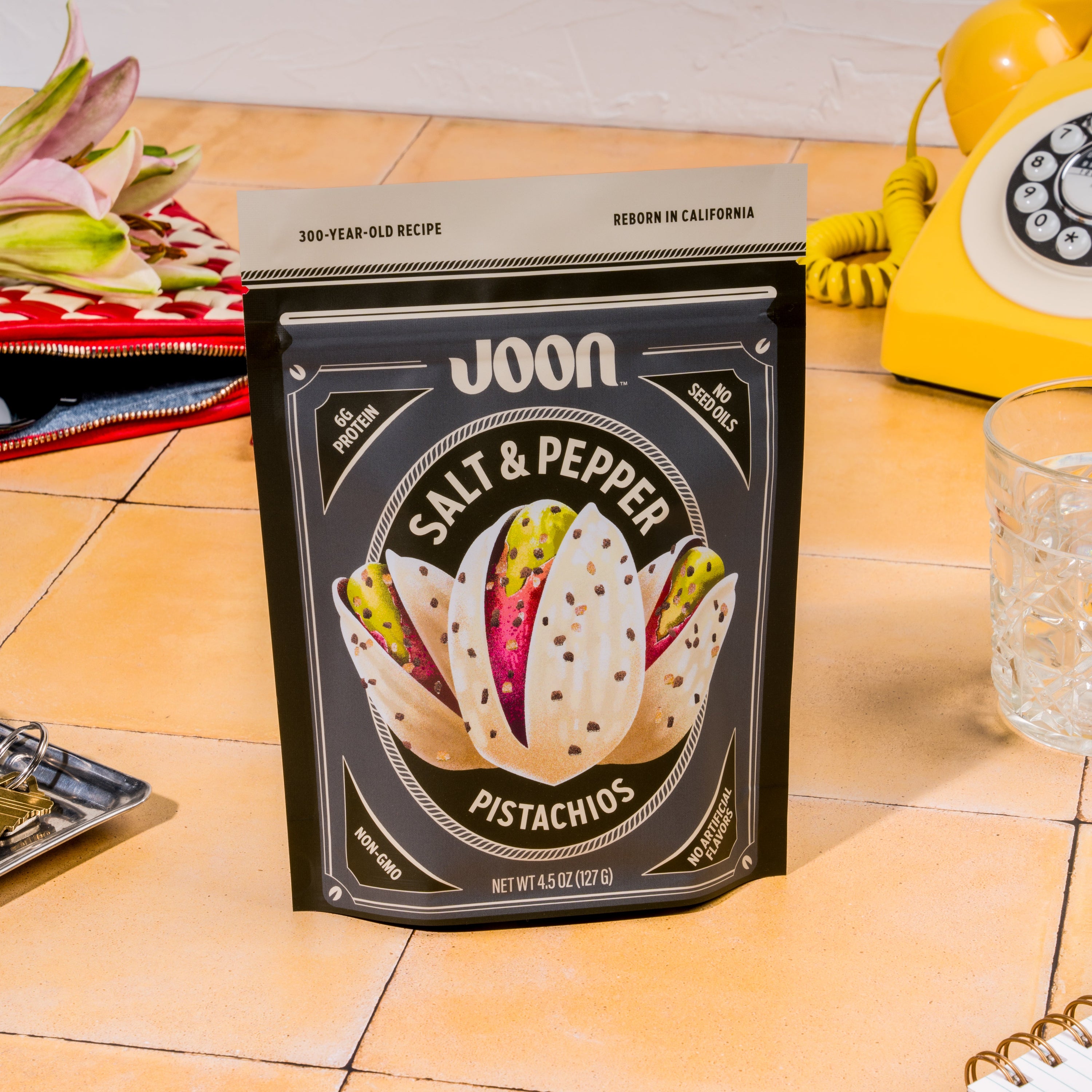 Joon Salt & Pepper Pistachios 4.5 oz bag on a yellow tiled counter beside a yellow rotary phone.
